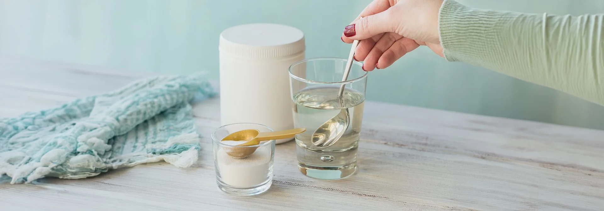 A hand stirring a scoop of collagen powder in a glass of water with the jar next to it.