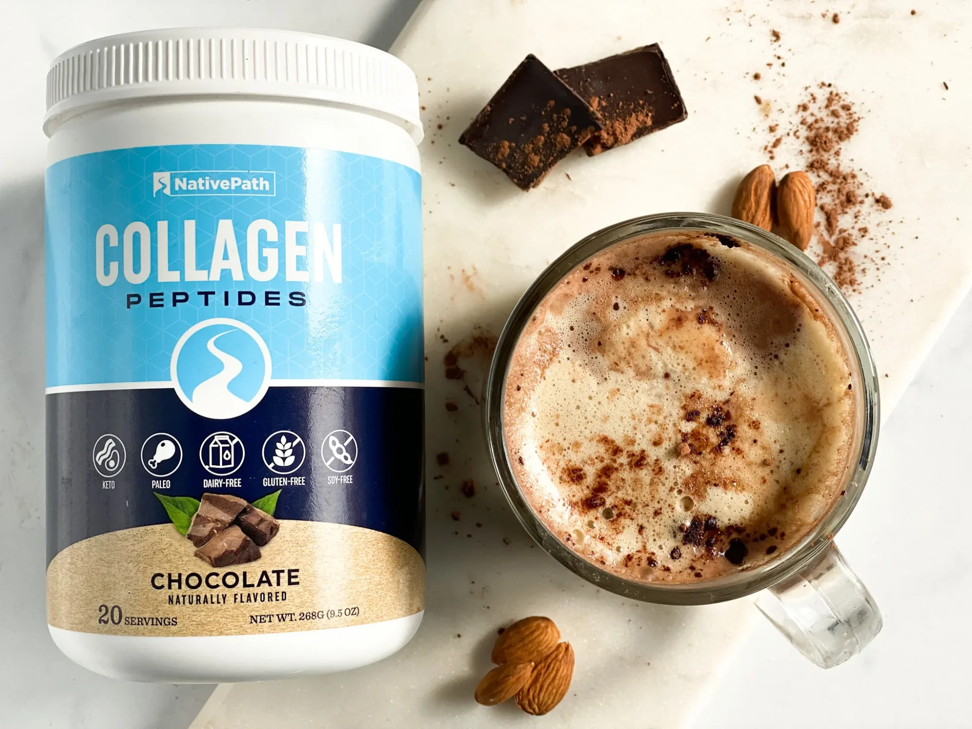 Top of view image showcasing a steaming mug of healthy hot chocolate with a jar of Chocolate Collagen. Almond, cocoa, and dark chocolate are sprinkled around the mug.