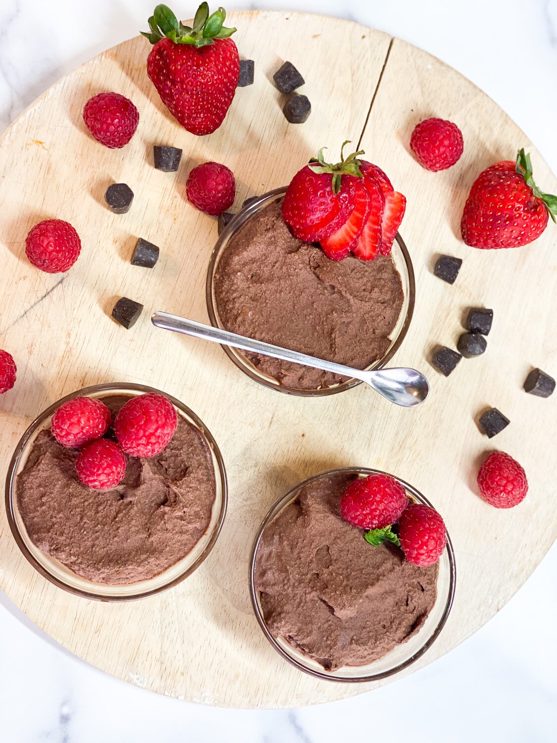 Overhead shot of three glass bowls on a wooden cutting board filled with chocolate mousse and topped with raspberries. 