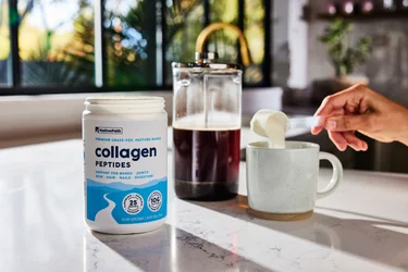 A hand pouring a scoop of NativePath Original Collagen Peptides from the container into a coffee mug with a French Press in the background.