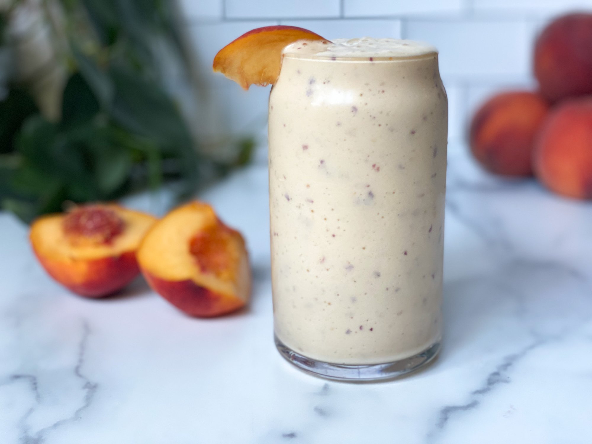 A peach smoothie in a class garnished with a slice of peach and peach halves in the background