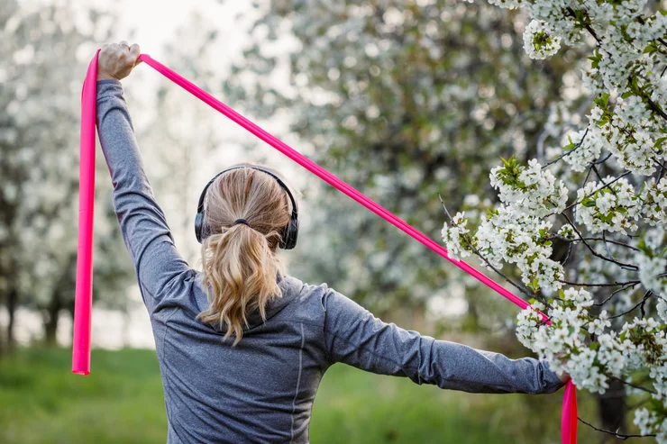 An older woman surrounded by white flowered trees stretching with a pink resistance band