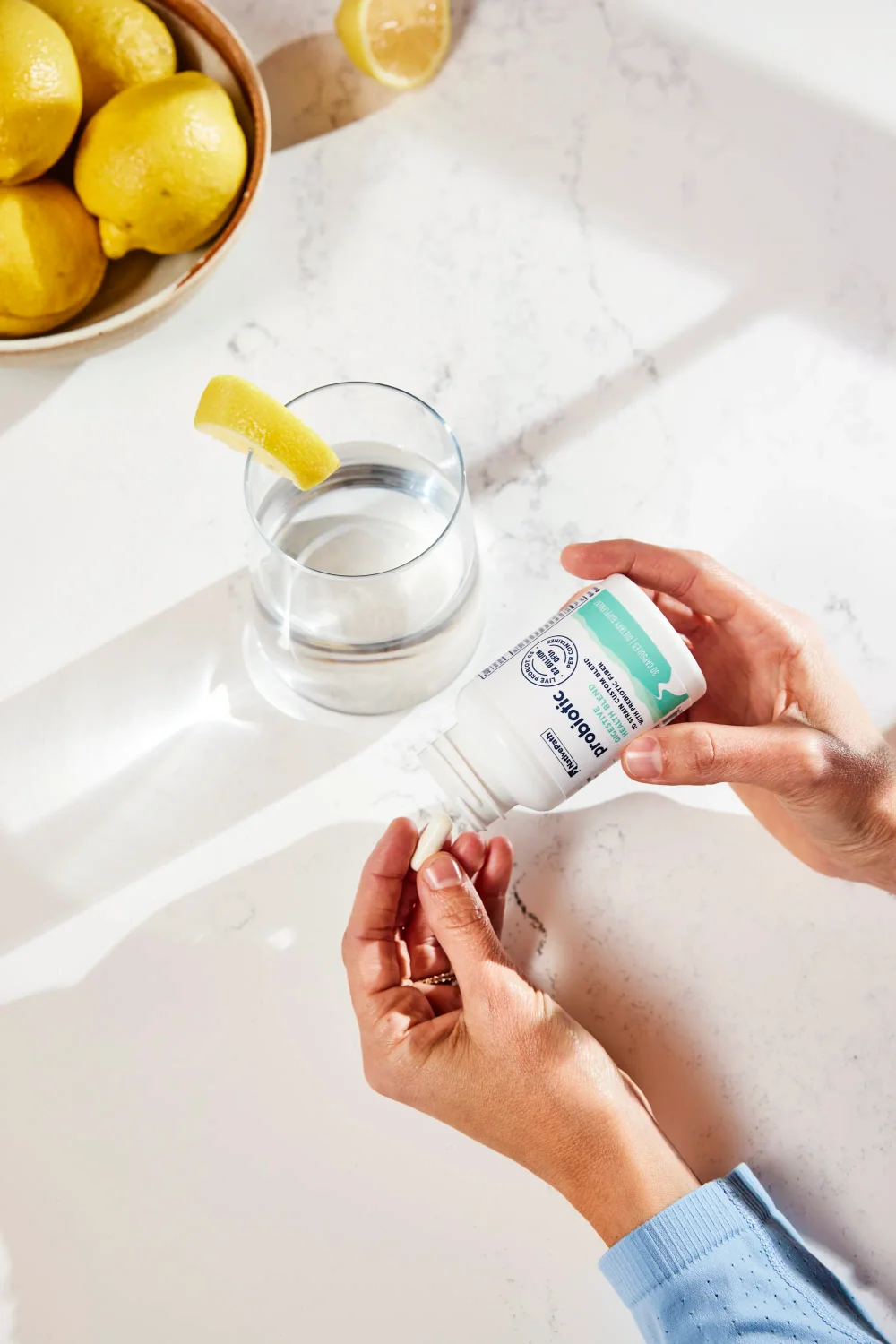 Top-view shot of a hand pouring out a NativePath Probiotic capsule into her other hand. Bowl of lemons and a glass of water in the background.