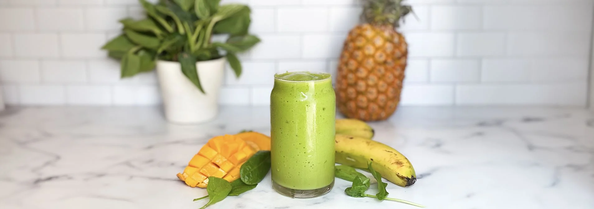 A clear glass filled with a green smoothie mixture. Mango, banana, and spinach surround glass. Pineapple and houseplant in background.