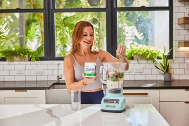 A woman pouring a scoop of NativePath Native Greens into a blender full of fruits and veggies