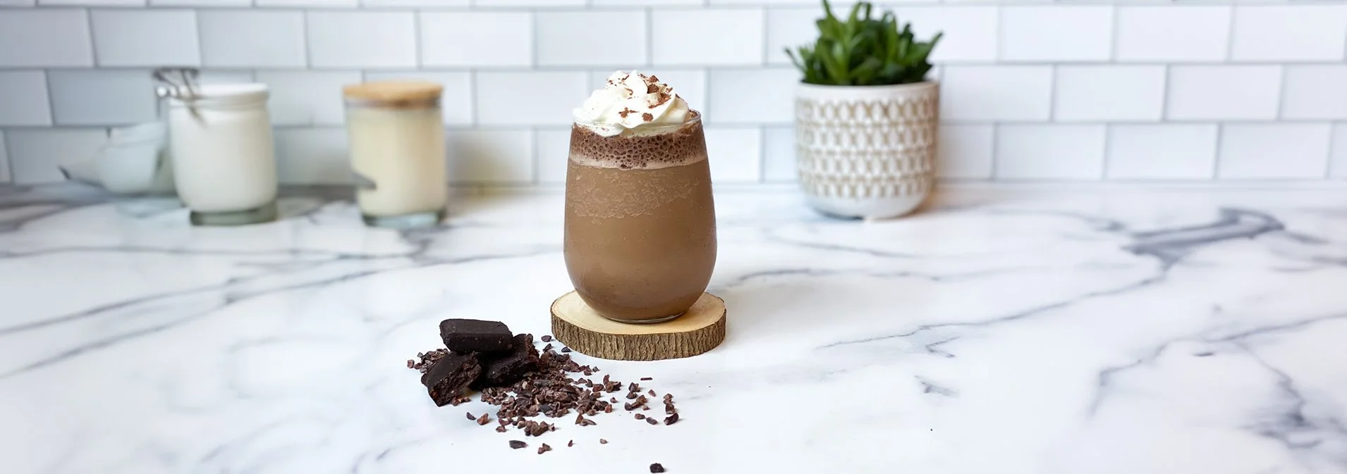 Front view of the Mocha Frappuccino in a glass cup on a wooden coaster with chocolate and cocoa nibs next to it and a plant in the background