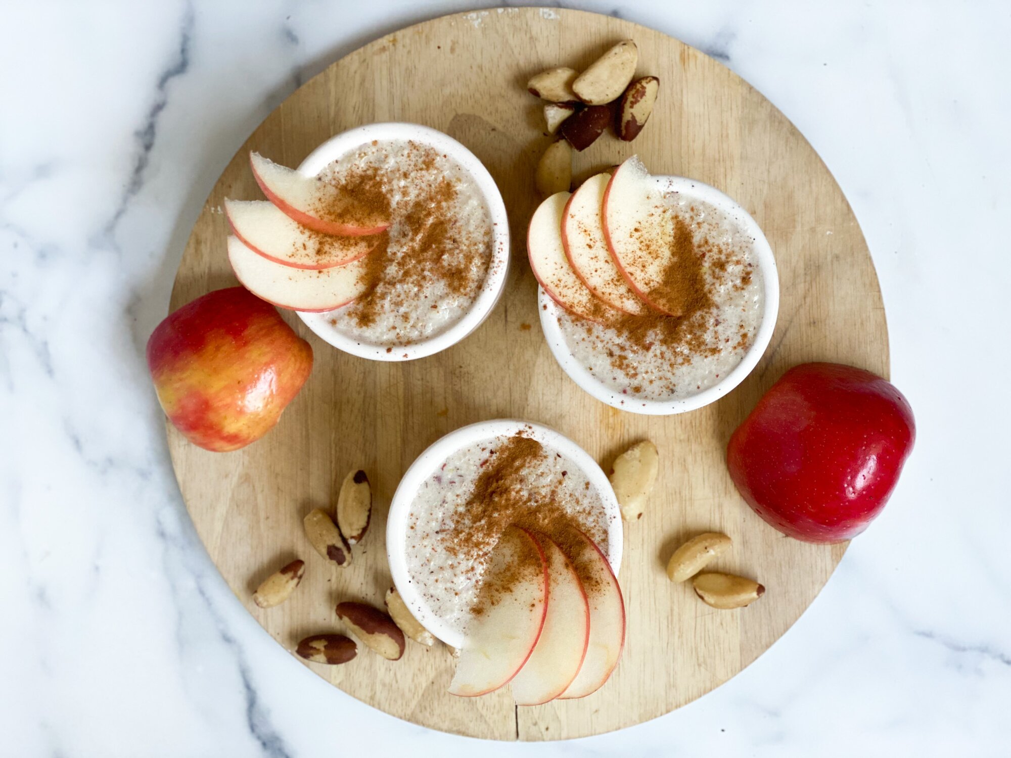 Top view of three ceramic bowls of porridge on a circular wooden board topped with apples and cinnamon.