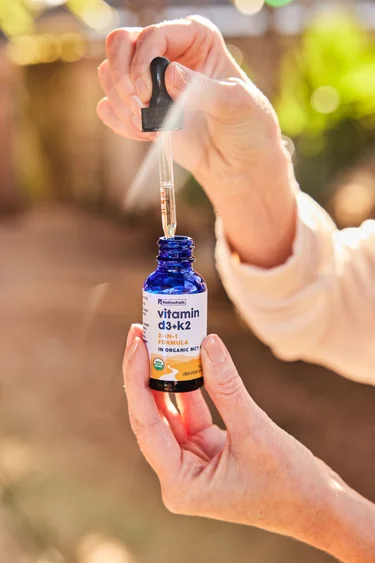 A woman's hands holding a bottle of NativePath Vitamin D3+K2 tincture outside with a sun flare