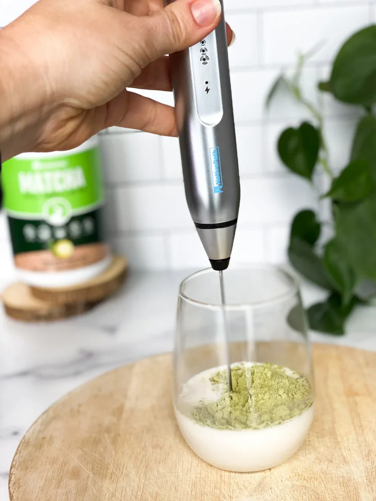 Woman frothing Matcha Collagen Powder into a clear glass of nut milk.