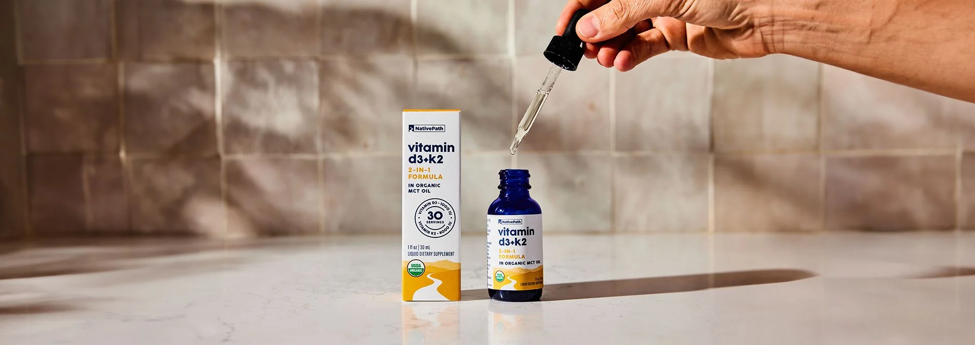 A bottle of NativePath Vitamin D3+K2 tincture with the dropper and packaging on a kitchen counter with a tile background
