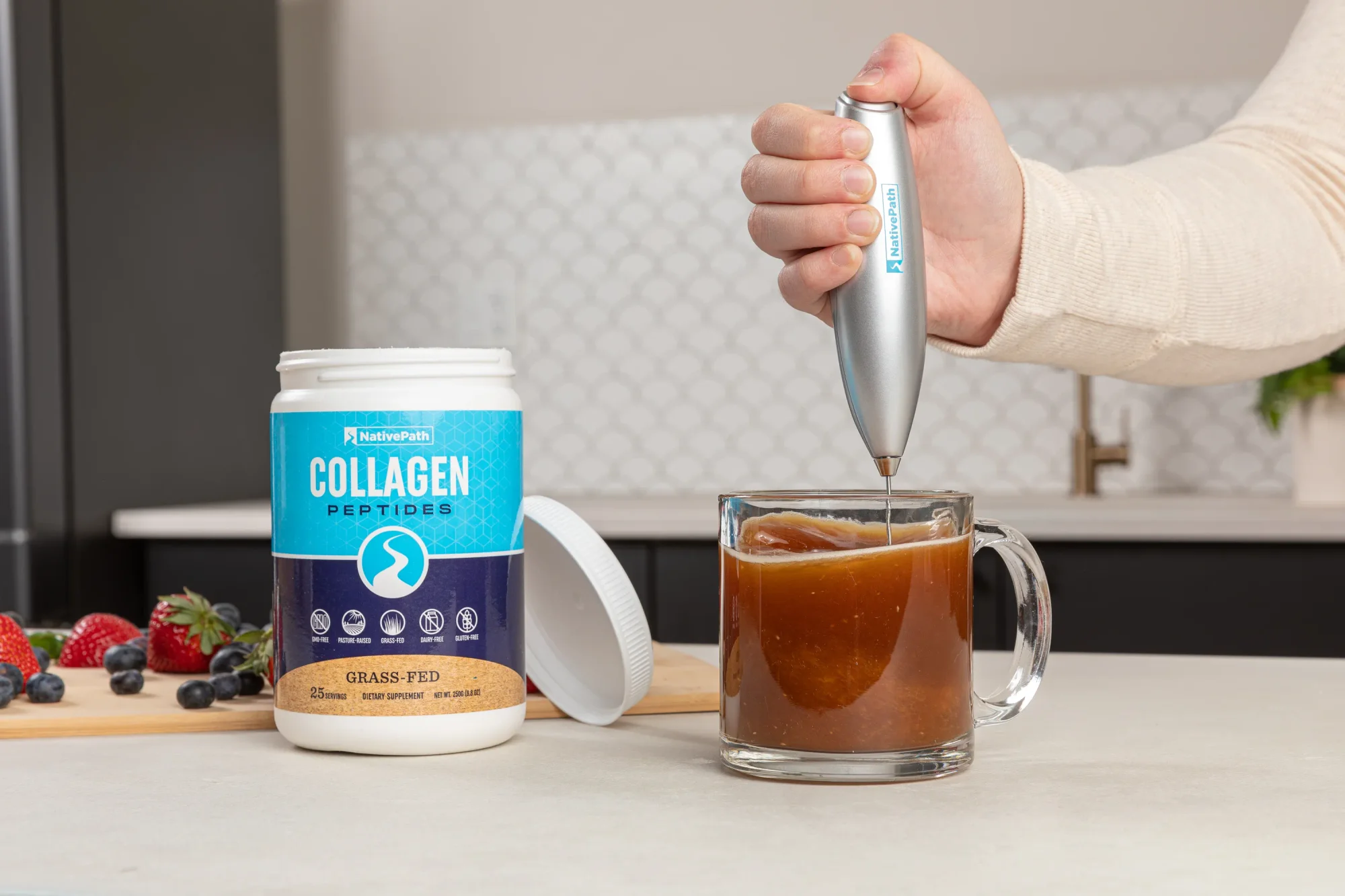 Hand holding a handheld frother. Frothing one scoop of collagen in a clear mug of coffee. NativePath Original Collagen jar off to the side. Kitchen setting.