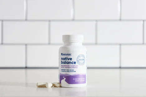 A bottle of NativePath Native Balance magnesium supplement with two capsules next to it on a counter with a white tile background
