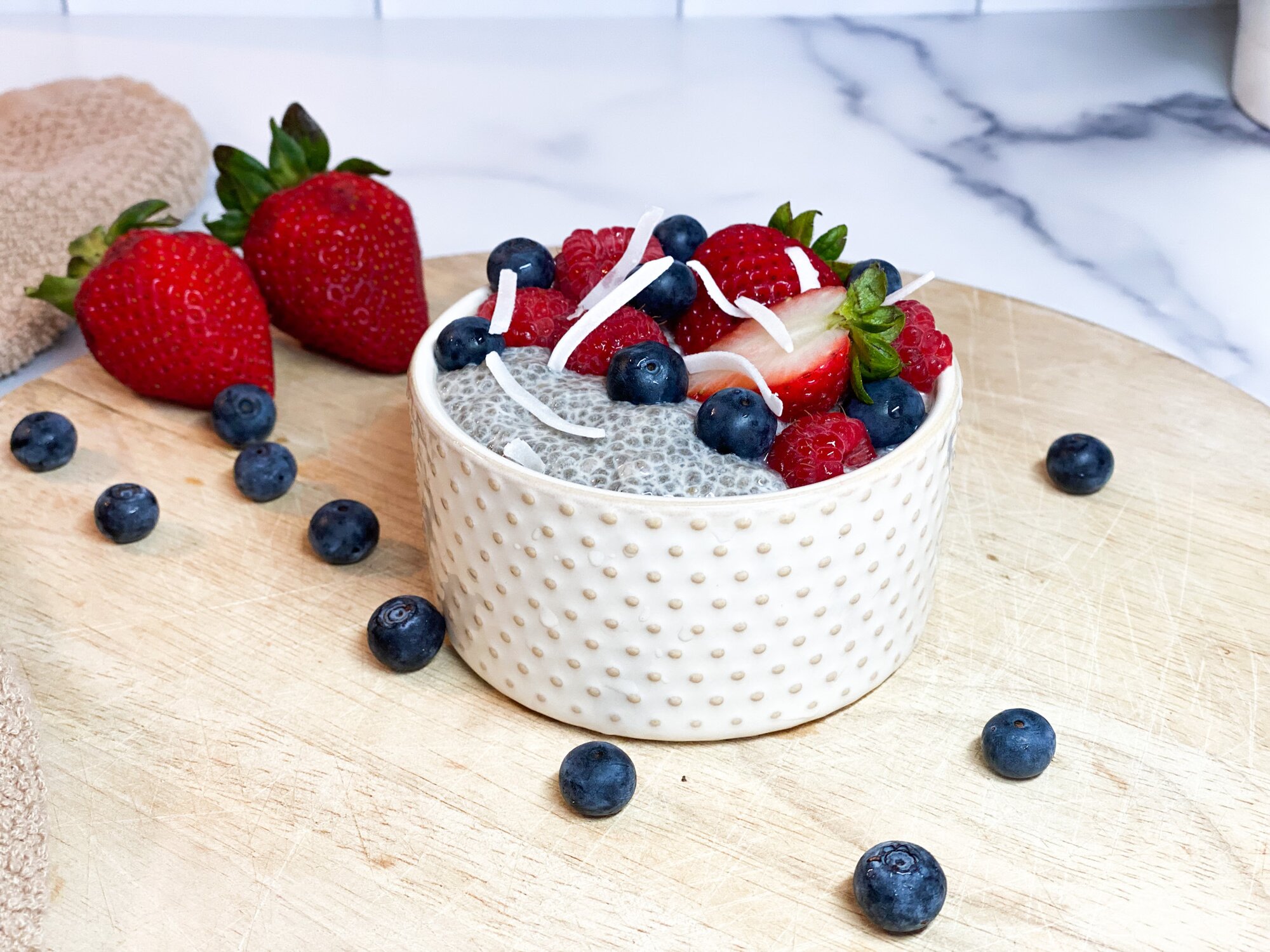 Ceramic bowl of vanilla chia pudding with fresh berries sprinkled on top. Strawberries laying next to bowl.