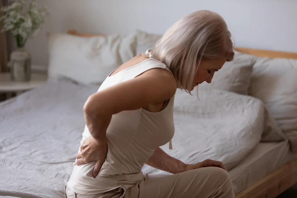 An older woman sitting on a bed with her hand on her lower back in pain.