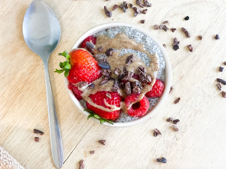 Ceramic bowl of vanilla chia pudding with nut butter, cocoa nibs, fresh berries sprinkled on top. Silver spoon laying next to bowl.