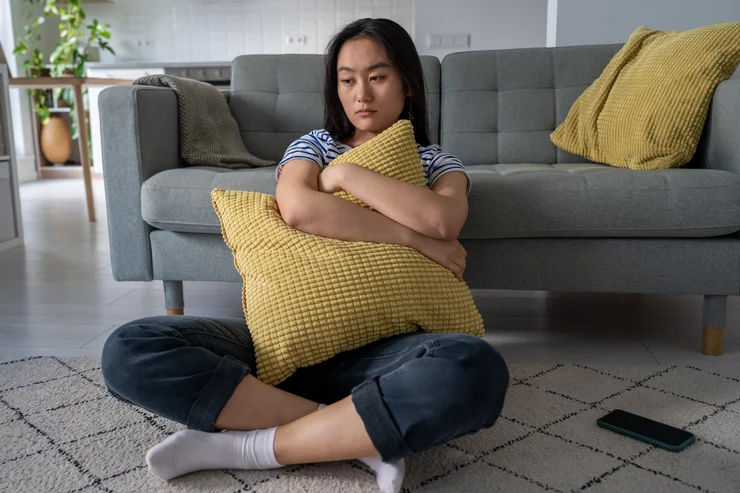 A sad looking woman sitting on the floor holding a pillow