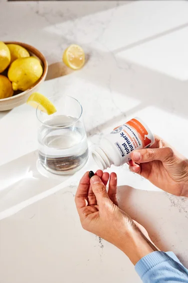 A hand pouring a softgel from a container of NativePath Total Turmeric with a glass of water and bowl of lemon in the background