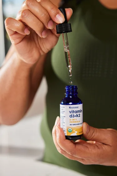 A close-up shot of a woman in a green shirt pulling a full dropper of NativePath Vitamin D3+K2 tincture from the bottle.