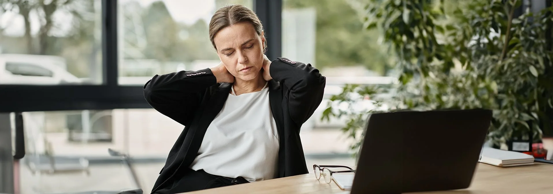 Older woman sitting at a desk with a laptop, holding her neck in pain.