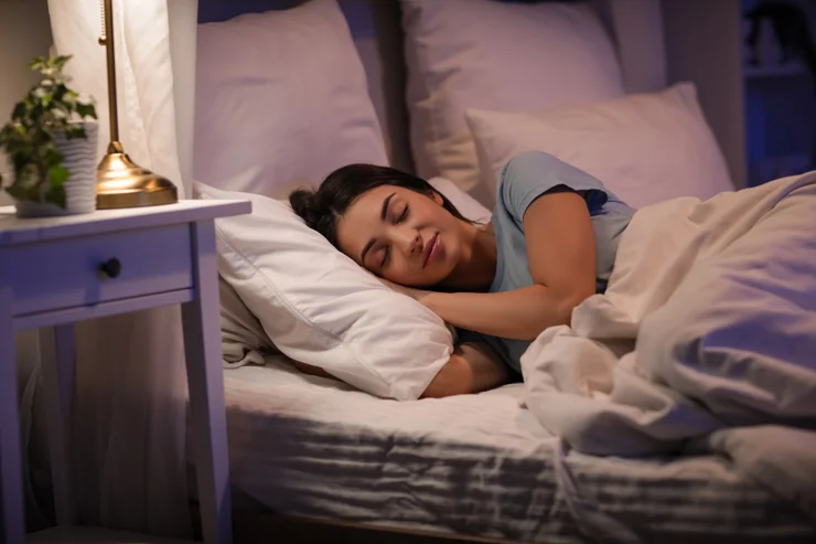 A woman asleep in a bed with a bedside lamp on next to her
