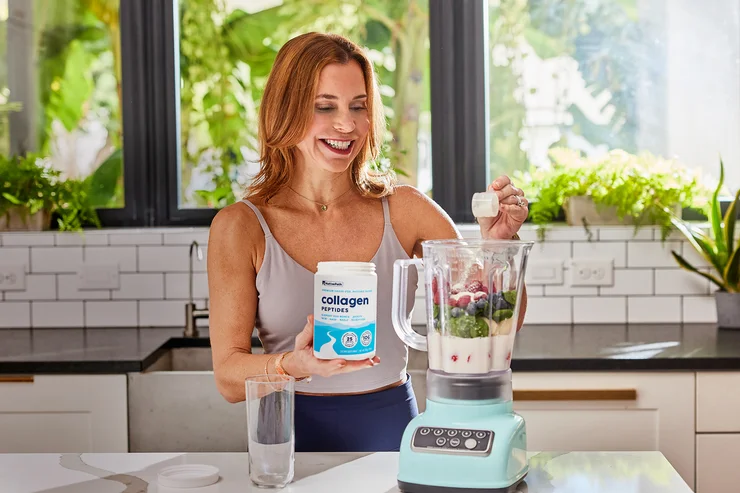A woman pouring a scoop of NativePath Original Collagen Peptides into a blender filled with fruits and veggies