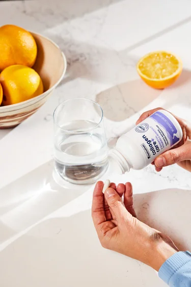 A woman pouring a capsule from a bottle of NativePath Collagen Care+ with water and lemons in the background