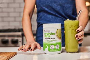 A woman's hand holding a glass of iced matcha with a container ofNativePath Matcha Latte next to it. 
