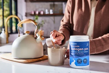 A woman pouring a scoop of NativePath Chocolate Collagen PM into a mug with a kettle next to her.