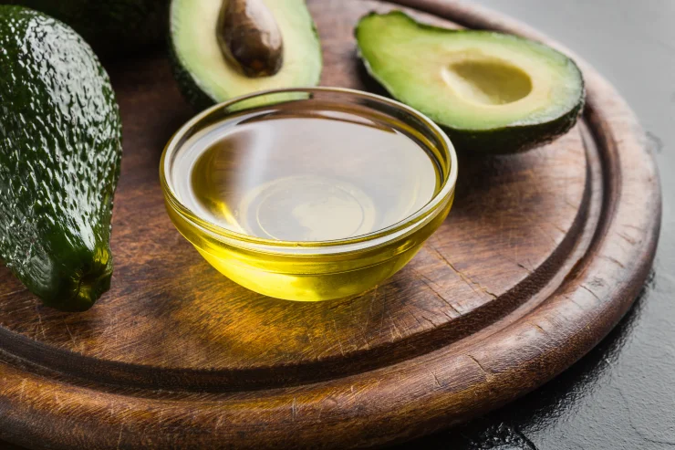 Avocado oil in clear bowl and fresh sliced avocadoes on wooden board.