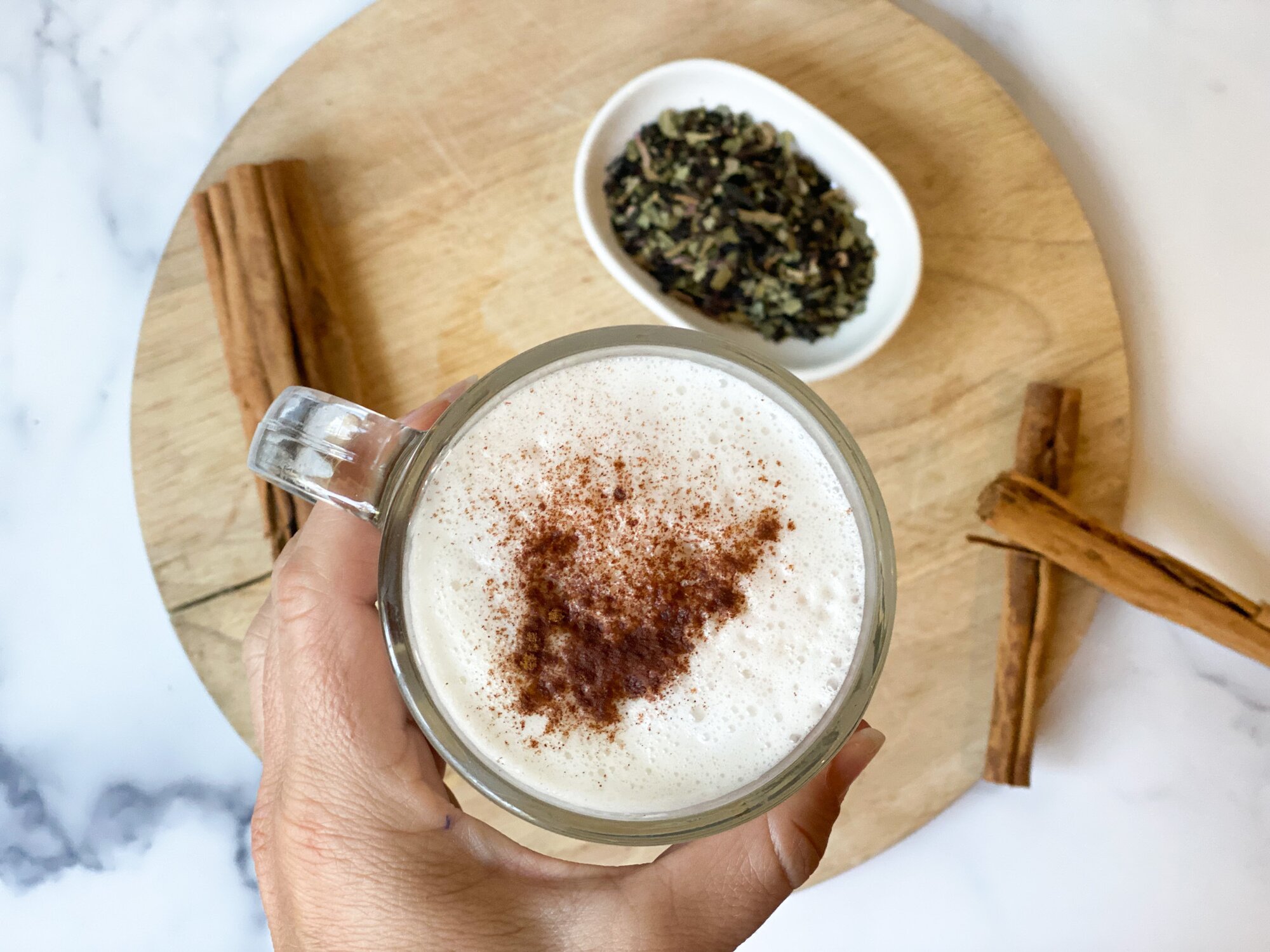 An overhead view of a hand holding a glass mug of vanilla chai latte with a wooden cutting board, bowl of chai tea, and cinnamon sticks in the background.