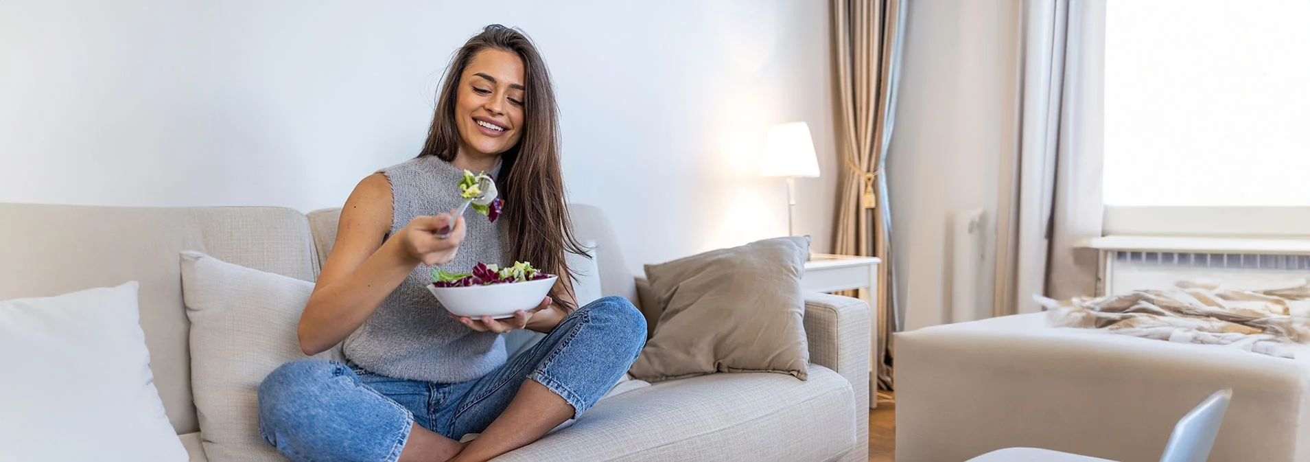 A woman sitting on a white couch eating a salad out of a bowl.