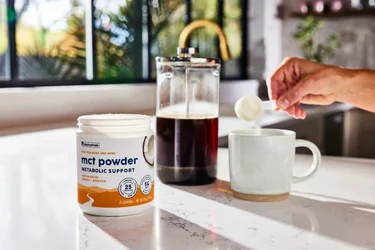 A hand pouring a scoop of NativePath MCT Powder into a coffee mug with a French Press in the background