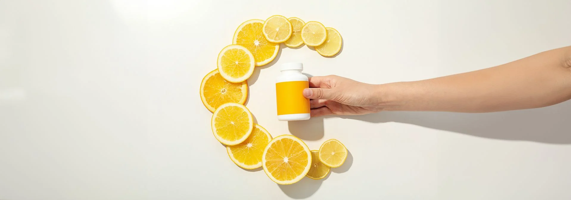 A hand holding a bottle of vitamin C supplements with the letter C made out of orange and lemon slices in the background