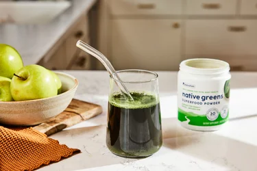 A glass full of NativePath Native Greens with the container and bowl of apples in teh background