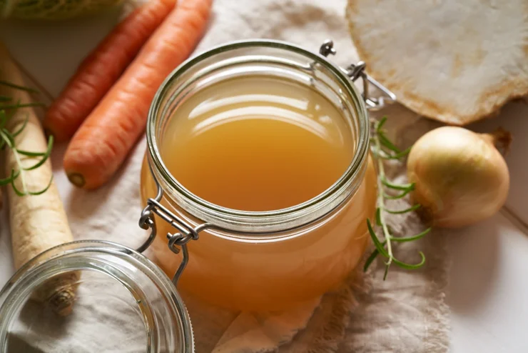 A glass jar of bone broth surrounded by carrots and onion 