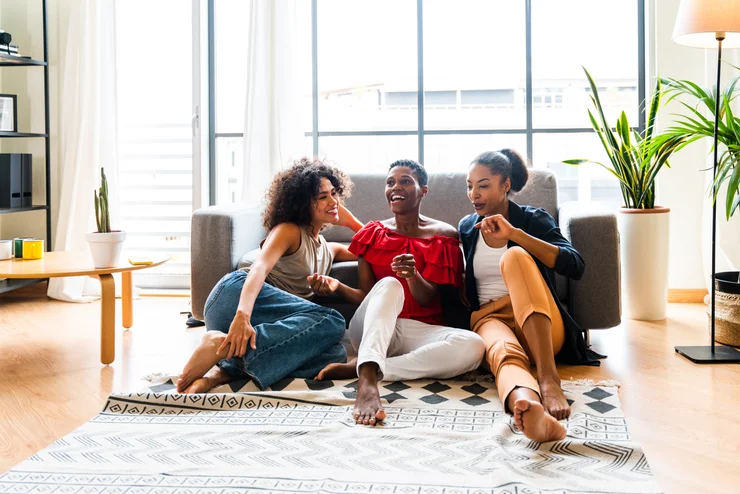 A group of femal friends sitting on the floor of a living room laughing.