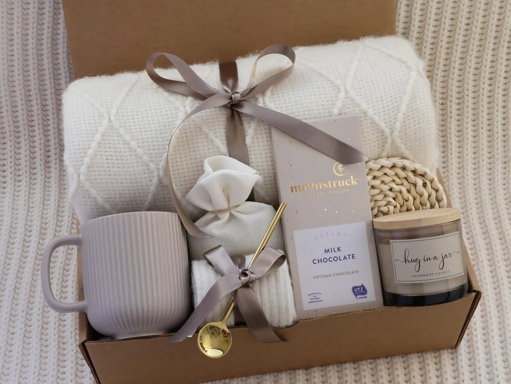 Gift box with a cream knit blanket, a brown mug, gold spoon, milk chocolate bar, candle labeled "hug in a jar," and a small wrapped item. Cozy and inviting.