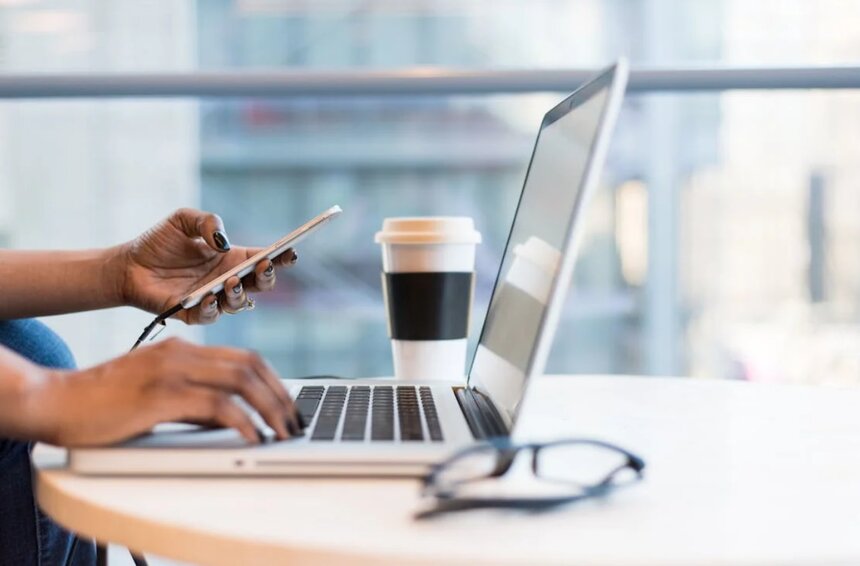 A woman sits at a desk, using her laptop while holding a cup of coffee, focused on her work.