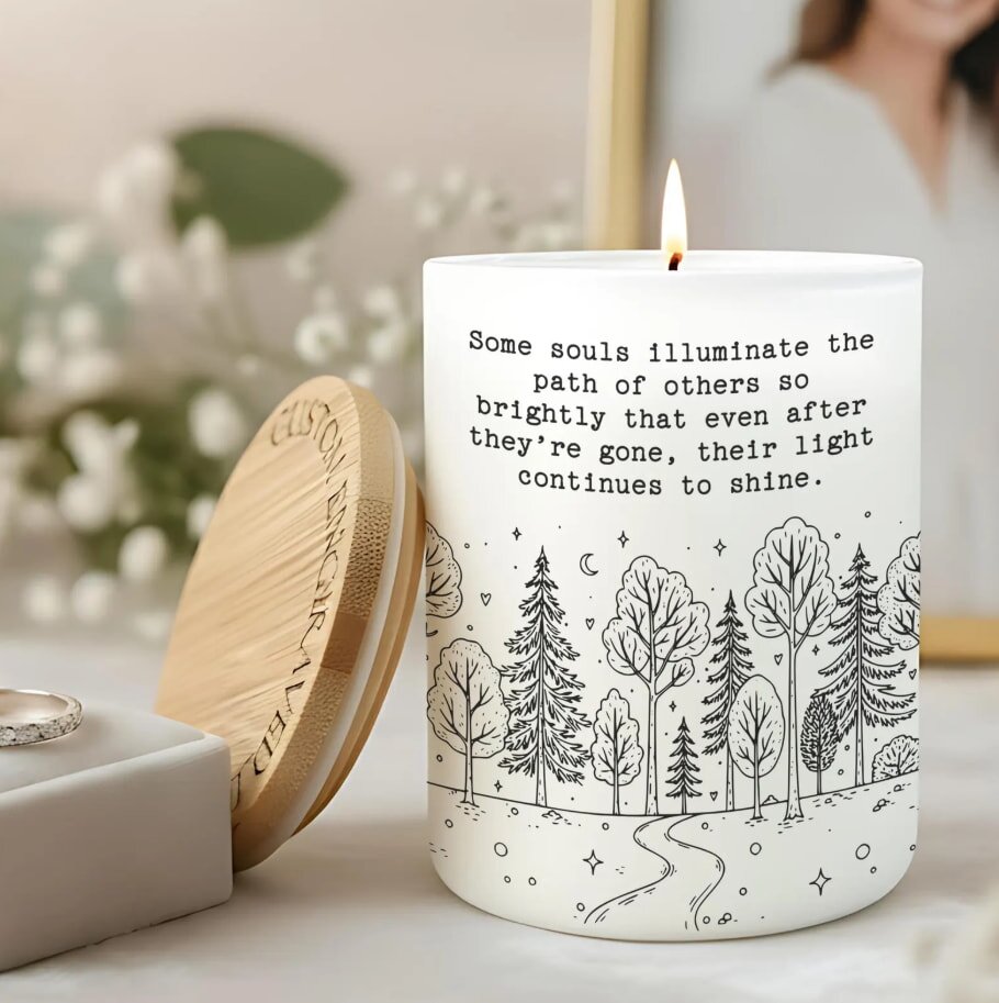 Lit candle with a decorative lid engraved "Custom Engraved," featuring a forest illustration and quote about souls shining on. Background has blurred flowers.