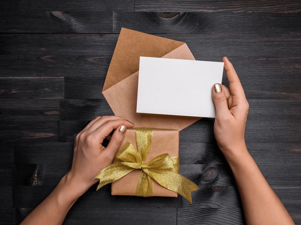 Hands holding a blank card and brown envelope above a gift wrapped in brown paper with a gold ribbon, on a dark wooden table.