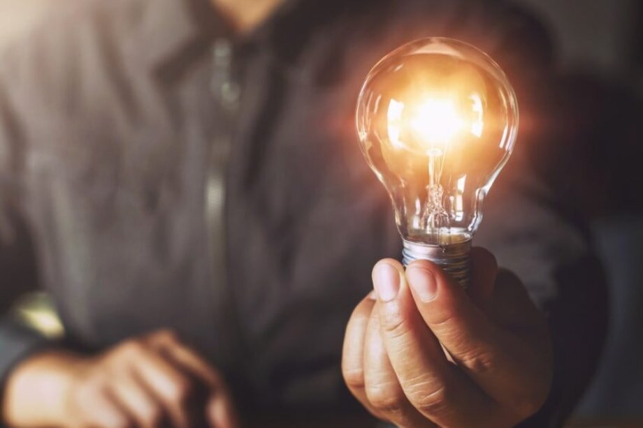 A person holds a glowing lightbulb in their hand, symbolizing creativity and ideas. The warm light contrasts with the dark, blurred background.