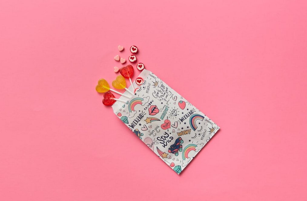 Colorful paper bag with doodles, including rainbows and hearts, on a pink background. Lollipops and candy hearts spill out, conveying a playful, cheerful vibe.