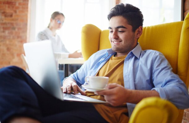 A man seated in a chair, working on a laptop while enjoying a cup of coffee.
