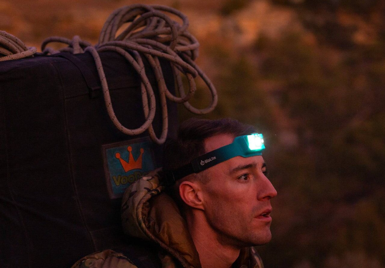 A man balances a backpack on his head, which has a light attached to it, creating a whimsical and unusual scene.