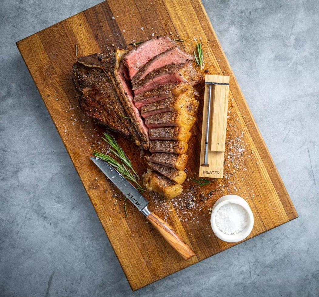 MEATER Pro wireless probe inserted in a steak on a grill, next to its charging block.