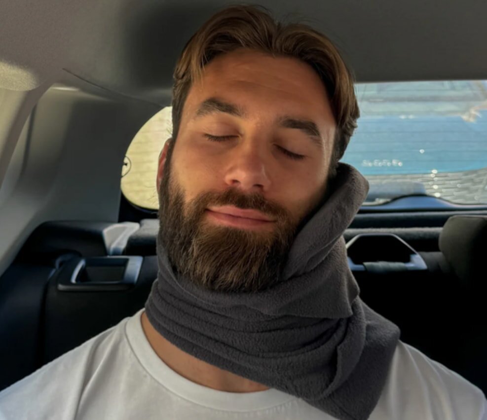 A bearded man wearing a neck pillow relaxes in the back seat of a car.