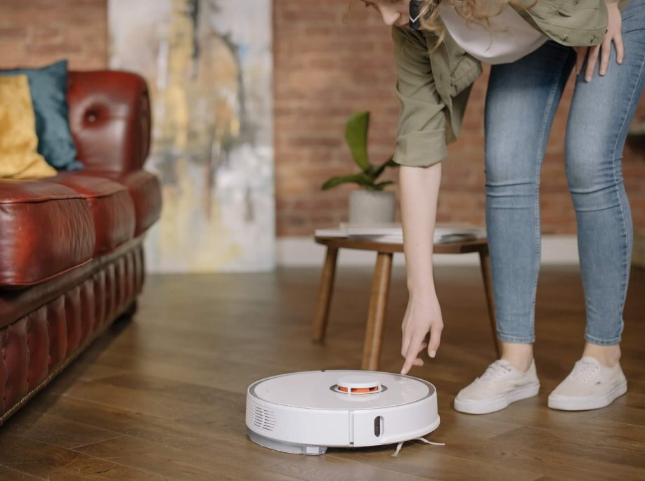 A person in jeans and white sneakers interacts with a white robotic vacuum on a wooden floor. The brick wall and red sofa create a cozy setting.