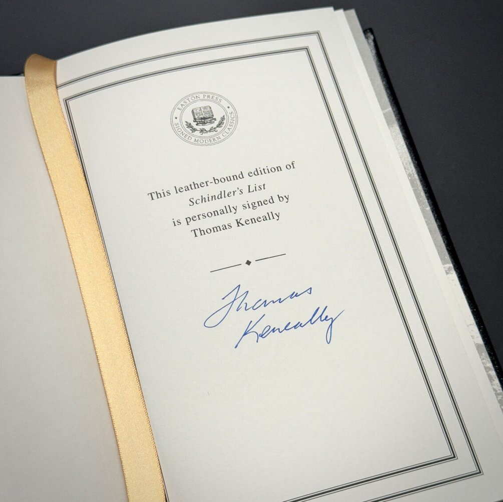 A page from a leather-bound book shows a gold ribbon and a printed note indicating that this edition of "Schindler's List" is signed by Thomas Keneally, with his signature in blue ink.
