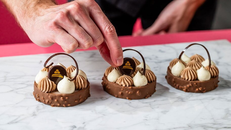 A hand skillfully places a chocolate decoration on one of three elegant desserts. Each features chocolate mousse swirls atop a nutty base on a marble surface.