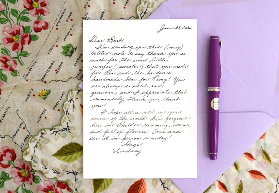 Handwritten letter on white paper with cursive script, placed on a floral map-themed fabric. A purple pen lies beside a lilac envelope, creating an elegant, heartfelt scene.
